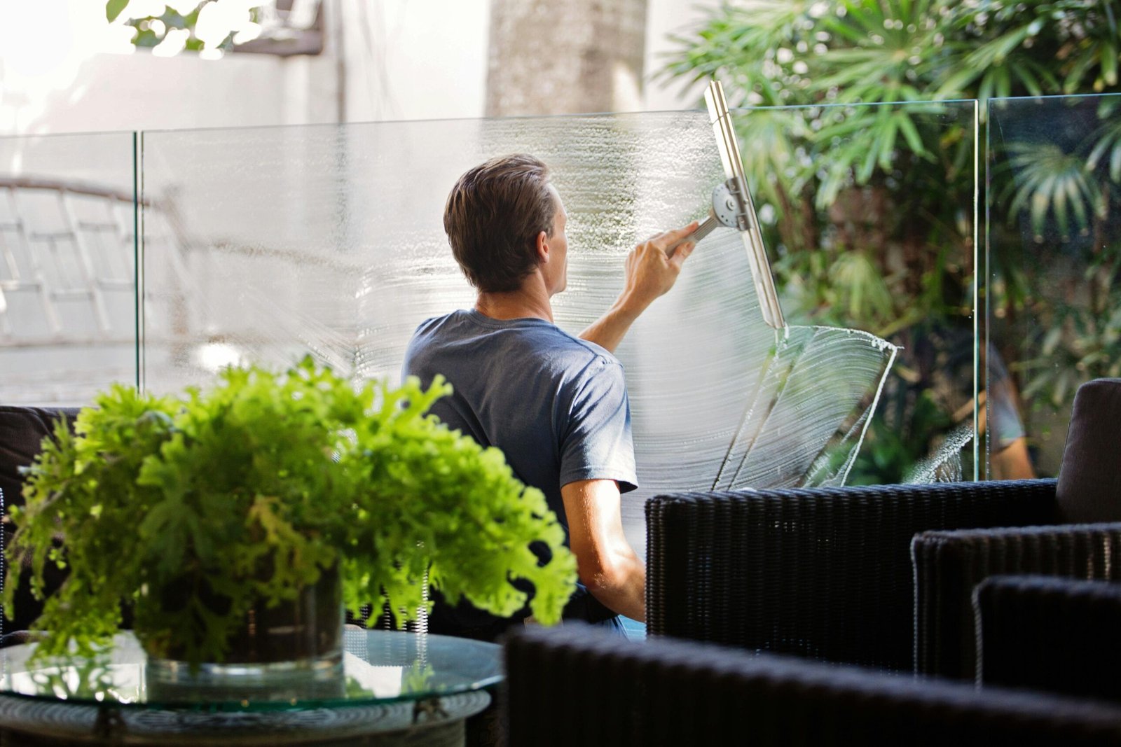A man cleaning a glass barrier outdoors with a squeegee, surrounded by greenery.